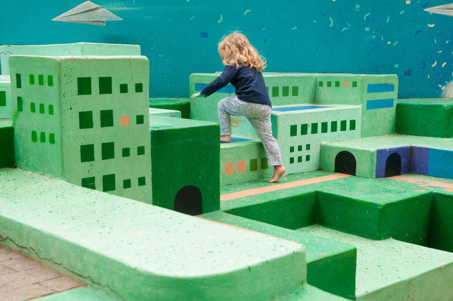 Foto de uma menina brincando em uma estrutura de cimento pintada de verde com vários blocos de alturas diferentes, cada um pintado como um prédio, uma casa, assemelhando-se a uma cidade, de modo que parece que a criança é um 'gigante subindo' pelos prédios de uma cidade em um dia ensolarado com uma parede ao fundo pintada de azul, como um grande céu.